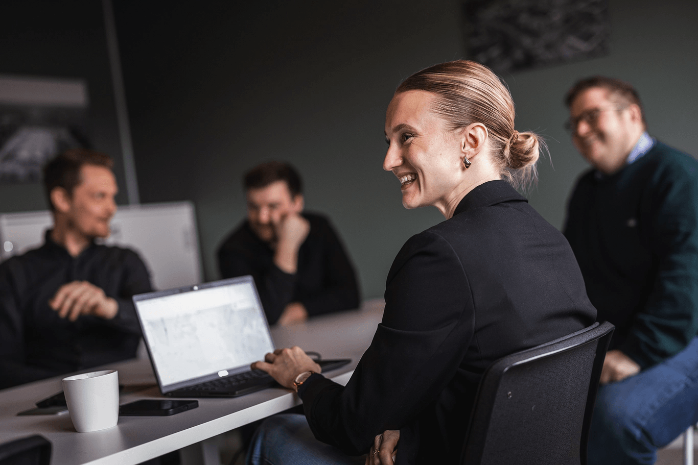 Kvinna med laptop sitter och ler vid ett mötesbord med tre kollegor i en avslappnad kontorsmiljö.
