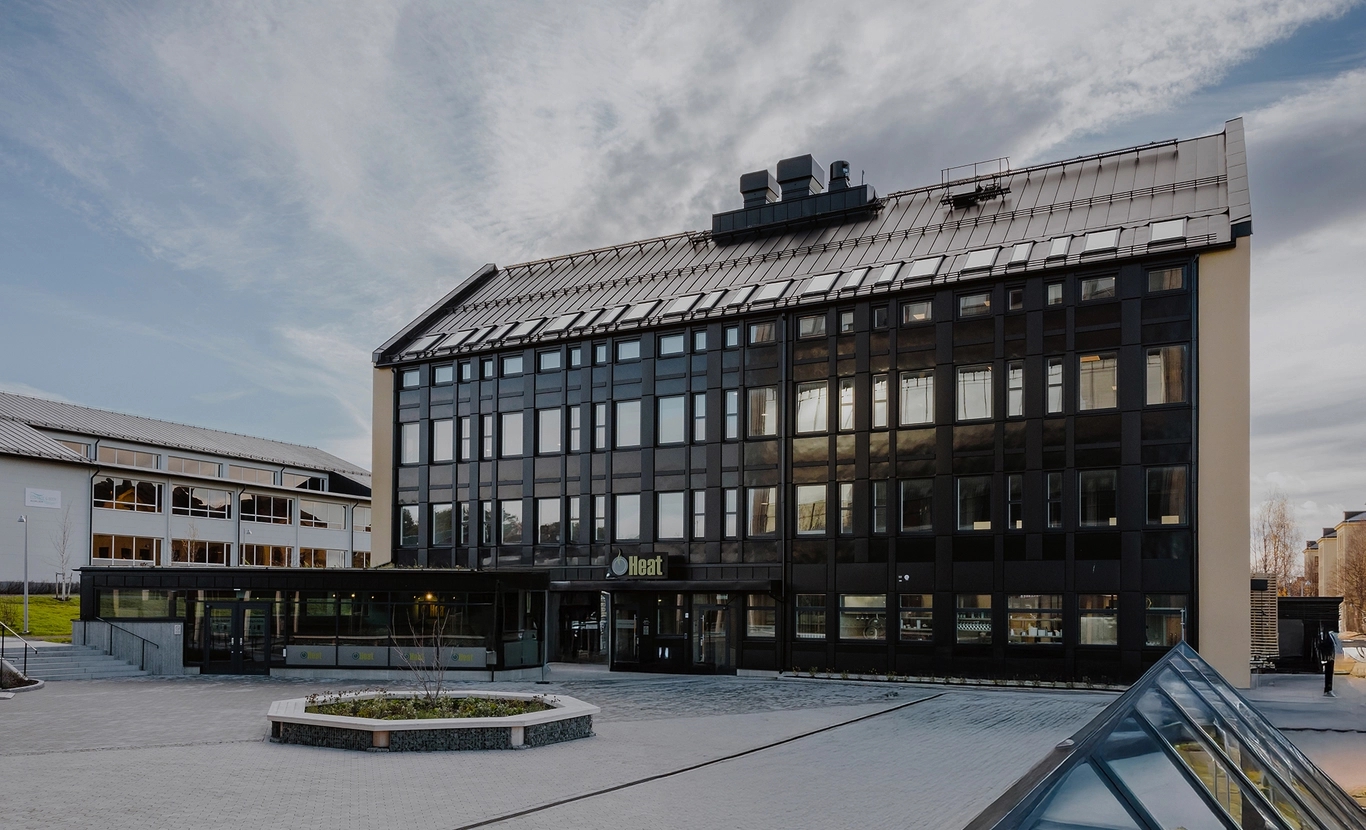 En modern, svart byggnad i fyra våningar med stora fönster och en innergård framför under en molnig himmel.