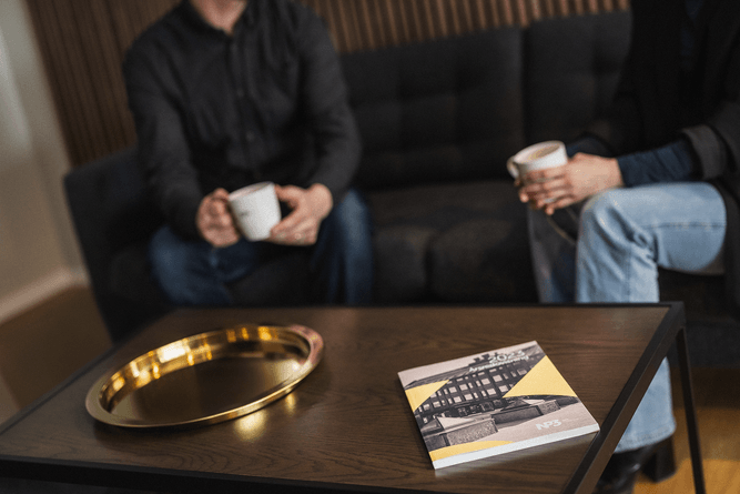 Två personer sitter i en mörk soffa med muggar i handen, med en bricka och en bok på ett soffbord av trä framför sig.