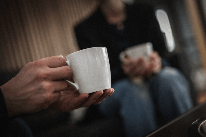 Två personer sitter tätt intill varandra med varsin vit mugg i handen, med fokus på händerna och muggarna.