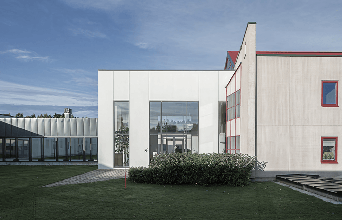 Modern tvåvåningsbyggnad med vit fasad, stora fönster och omgivande grönska under en delvis molnig himmel.